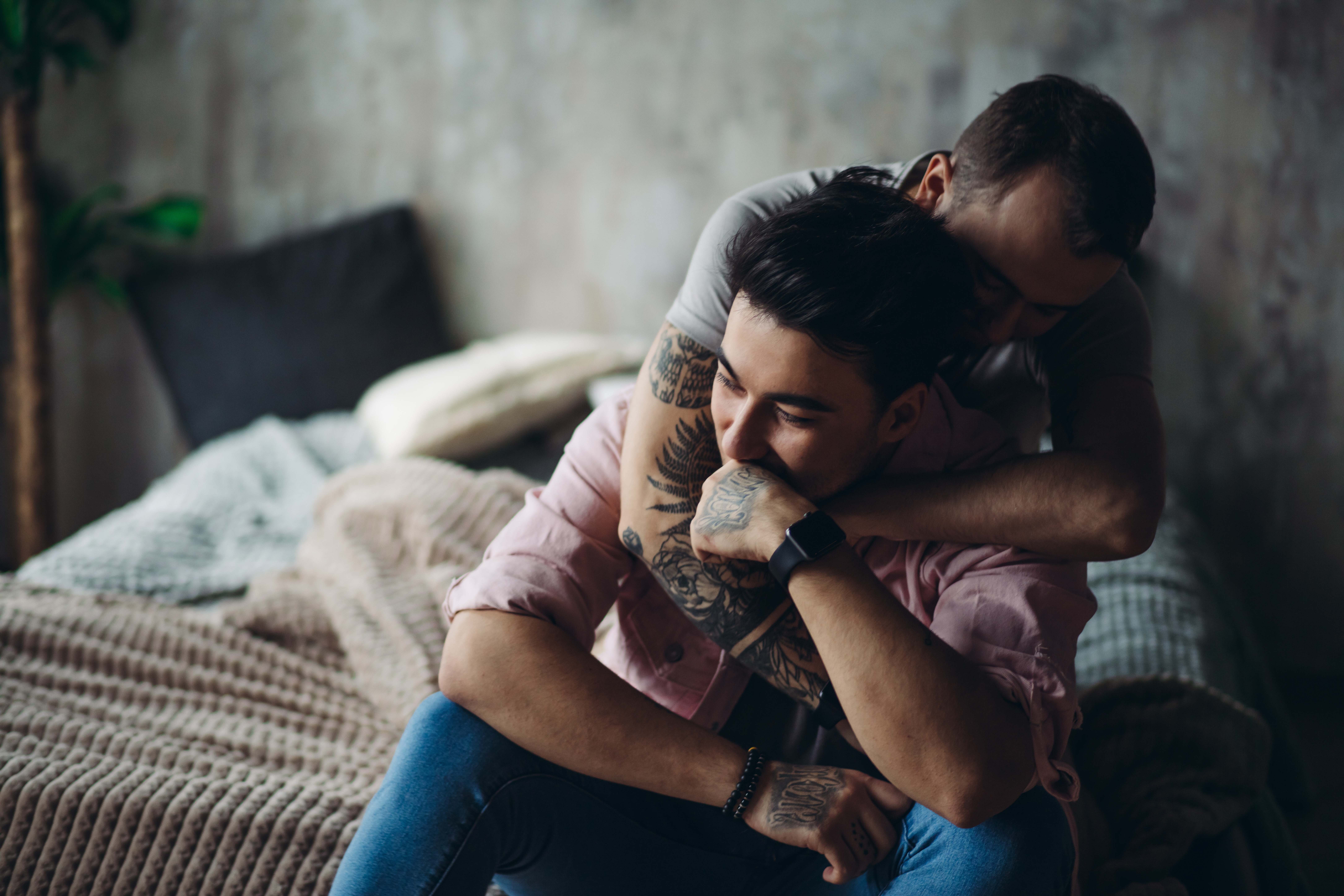 Happy gay couple embraced, joking and having fun in an intimate hug. Positive blue man embraces his friend, happy to be close to lover, sitting on bed at home, close up. Same-sex love relationship.