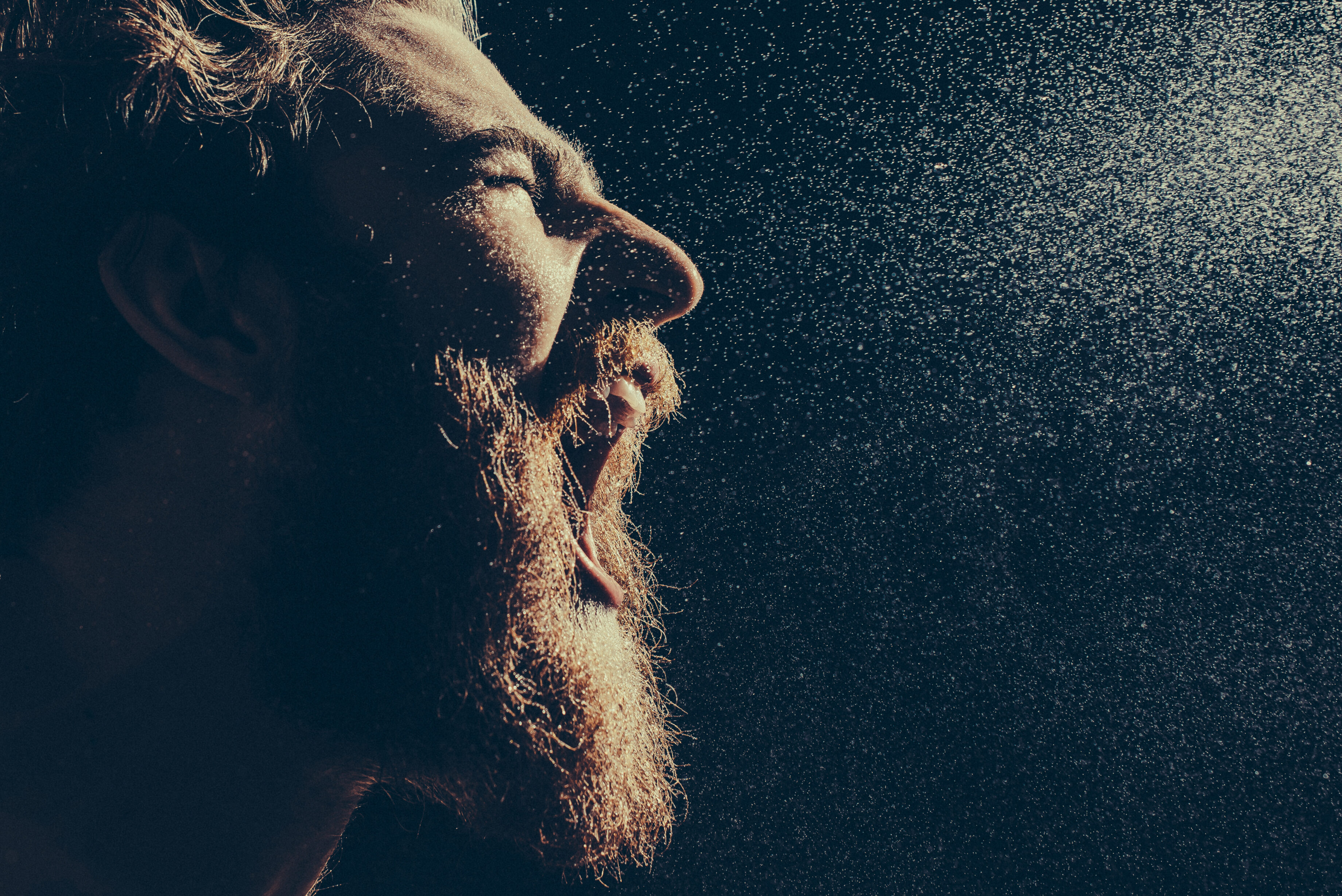 Naked bearded man angrily screams into a spray of water against a black background with copy space. Emotional portrait of a man like a barbarian. Toned image. side view close up view